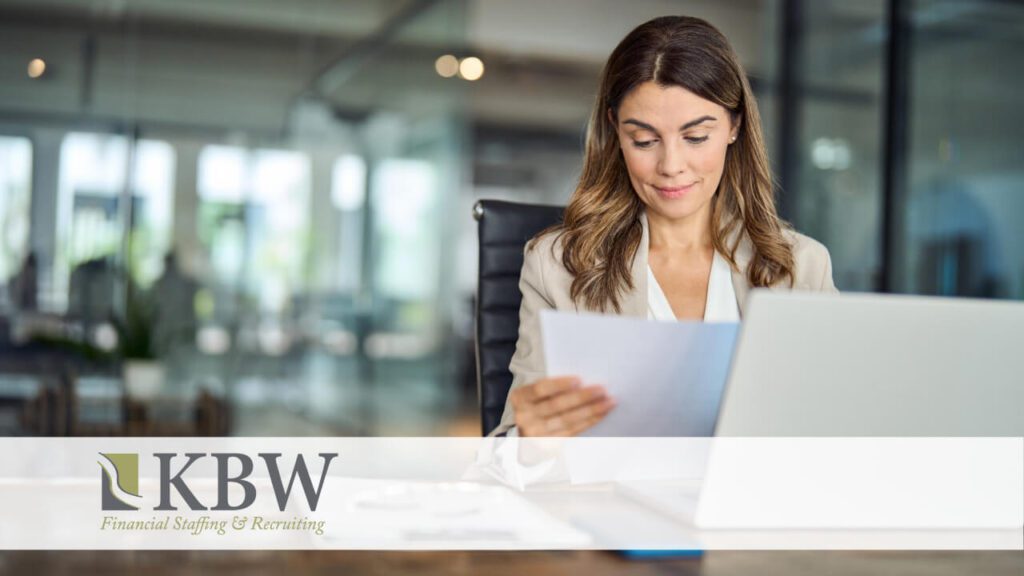 A business woman in an office sitting at her laptop looking down at a paper she's holding